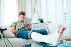 Junger Mann mit gebrochenem Bein sitzt auf dem Sofa und bedient ein Tablet.