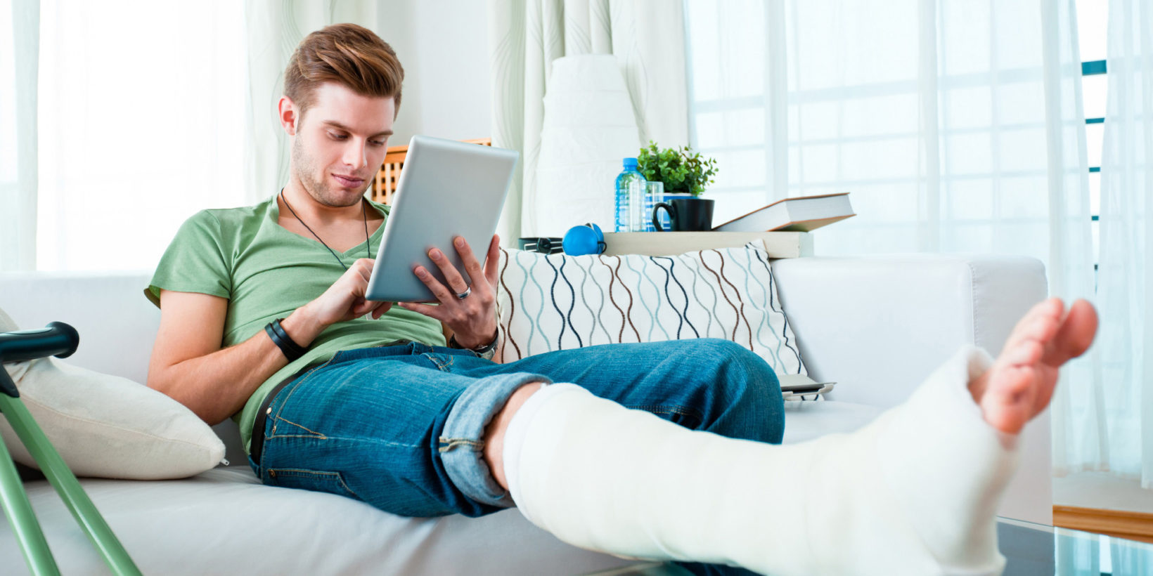 Junger Mann mit gebrochenem Bein sitzt auf dem Sofa und bedient ein Tablet.